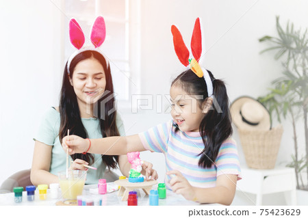 Attractive young woman with little cute girl are preparing for Easter celebration. Mom and daughter wearing bunny ears are spending time together before Easter while painting eggs. Attractive young woman with little cute girl are preparing for Easter celebration. Mom and daughter wearing bunny ears are spending time together before Easter while painting eggs. 75423629