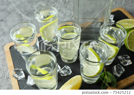 Board with bottle and shots of vodka with lime on gray background Board with bottle and shots of vodka with lime on gray background 75425212