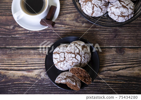 Chocolate cookies with cracks, strewed with icing sugar. Homemade baking, wooden background. Background: a cup of coffee and a plate of cookies. Selective focus, close-up Chocolate cookies with cracks, strewed with icing sugar. Homemade baking, wooden background. Background: a cup of coffee and a plate of cookies. Selective focus, close-up 75434280