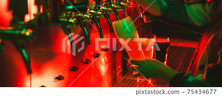 Young woman barman pouring a cold lager beer from tap to glass in neon light. Young woman barman pouring a cold lager beer from tap to glass in neon light. 75434677