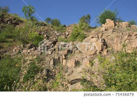 A rocky slope of an abandoned granite quarry in the Nikolaev region of Ukraine on a hot summer day. Sharp stones overgrown with sparse vegetation. Blue cloudless sky over the cliffs. 75436879