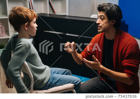 Side view of focused schoolboy sitting on chair listening young music teacher talking and gesturing 75437016