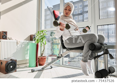 Young attractive woman in hijab, performs exercises with dumbbells, sitting on a bench at home gym. 75437526