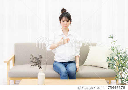 一個年輕女子坐在窗邊的沙發上,把她的手放在她的胸部 一個年輕女子坐在窗邊的沙發上,把她的手放在她的胸部 75446676