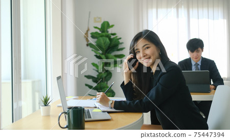 Happy businesswoman working with computer laptop and talking on mobile phone while sitting with her colleague in modern office. Happy businesswoman working with computer laptop and talking on mobile phone while sitting with her colleague in modern office. 75447394