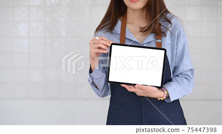 Cropped shot of young woman coffee shop owner in apron showing digital tablet. Blank screen for advertising text. Cropped shot of young woman coffee shop owner in apron showing digital tablet. Blank screen for advertising text. 75447398