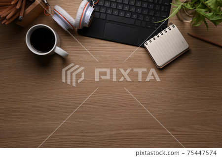 Top view of office desk with coffee cup, keyboard, headphone, plant, notebook and copy space on wooden background. Top view of office desk with coffee cup, keyboard, headphone, plant, notebook and copy space on wooden background. 75447574