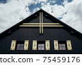 Gable roof of cottage with cloudy sky in background.There are 3 glass windows with wooden decorated shutters. Gable is made of wooden planks with dominant wooden cross at its top. 75459176