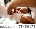 Close-up of male hands (3 hands) fastening cufflink at sleeve of white shirt in helping each other. The scene is in bright light with shallow depth of field. Focused on the cufflink. 75459178