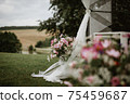 Flower pot full of different types of flowers in bloom, next to it is wedding altar with white curtains on grassy meadow. In background blurred hills, forest and lake. In foreground part of flowers. 75459687