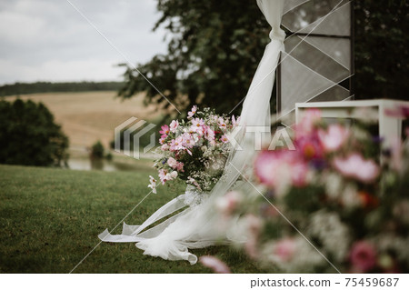 Flower pot full of different types of flowers in bloom, next to it is wedding altar with white curtains on grassy meadow. In background blurred hills, forest and lake. In foreground part of flowers. 75459687