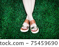A pair of human legs (female) with shoes lying crosswise in soft, deep green grass. Shoes are lightweight and open with leather straps and higher platform. A person has painted nails with red varnish. 75459690