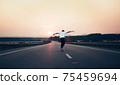 Man with arms outstretched riding a skateboard on the motorway road toward the setting sun in the background. The background is slightly blurred, focus on a skateboarder in the foreground. 75459694
