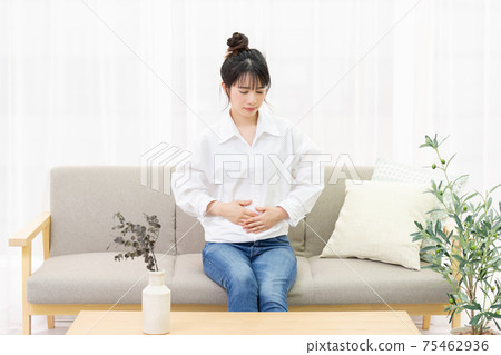 A young woman sitting on a sofa by the window and holding her belly by hand 75462936