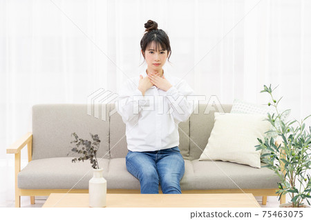 A young woman sitting on a sofa by the window and holding her throat 75463075