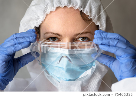 Female doctor in PPE suit close-up, nurse puts on medical glasses during COVID-19 pandemic Female doctor in PPE suit close-up, nurse puts on medical glasses during COVID-19 pandemic 75465355