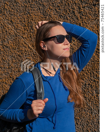 Young attractive woman in blue hoody black sunglasses on wall background listening to music with Wi-Fi headphones. 75465421
