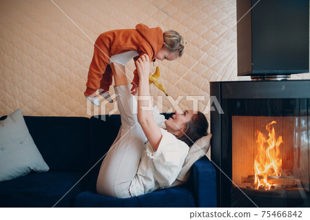 Mother and child sitting and playing on sofa near fireplace. Mom and baby. Parent and little kid relaxing at home. Family having fun together. 75466842