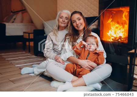 Mother and child sitting and playing on sofa near fireplace. Mom and baby. Parent with daughter and grandson little kid relaxing at home. Family having fun together. 75466843