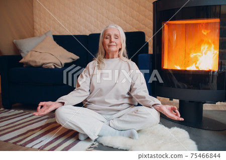 Portrait of senior woman sitting in lotus position indoors with fireplace. Yoga and meditation zen like concept. Portrait of senior woman sitting in lotus position indoors with fireplace. Yoga and meditation zen like concept. 75466844