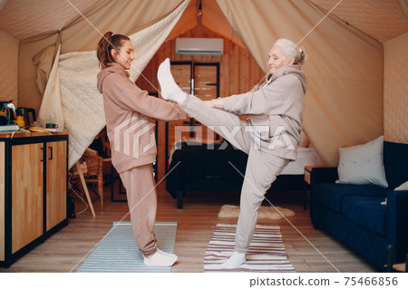 Family doing warming up exercises sports indoors. Young and senior elderly woman relaxing at glamping camping tent. Mother and daughter modern at fitness vacation lifestyle concept. Family doing warming up exercises sports indoors. Young and senior elderly woman relaxing at glamping camping tent. Mother and daughter modern at fitness vacation lifestyle concept. 75466856
