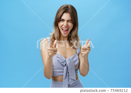 Charismatic joyful good-looking woman in trendy matching outfit winking joyfully making finger fun gesture at camera smiling broadly showing cute gapped teeth posing happy over blue wall 75469212