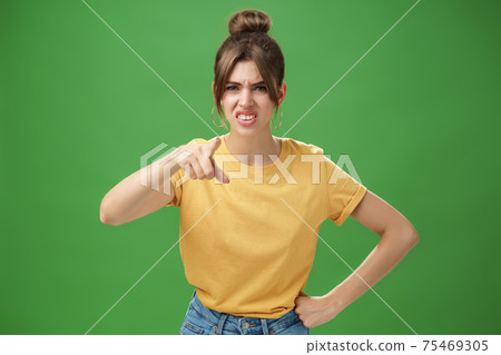 Portrait of arrogant impolite young woman in yellow t-shirt pointing at camera squinting and grimacing from disdain and scorn standing unimpressed judging against green background Portrait of arrogant impolite young woman in yellow t-shirt pointing at camera squinting and grimacing from disdain and scorn standing unimpressed judging against green background 75469305