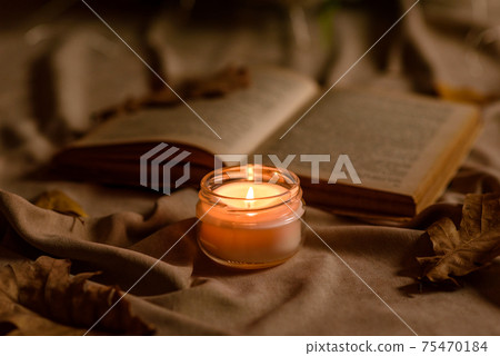 A burning candle on a wooden table in front of a book in a half-mast A burning candle on a wooden table in front of a book in a half-mast 75470184