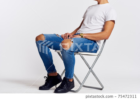 a man in jeans and a t-shirt sits on a chair on a light background side view a man in jeans and a t-shirt sits on a chair on a light background side view 75472311