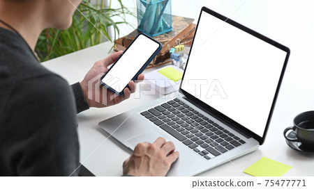 Cropped shot of young man designer using smart phone and working with computer laptop at workplace. Cropped shot of young man designer using smart phone and working with computer laptop at workplace. 75477771