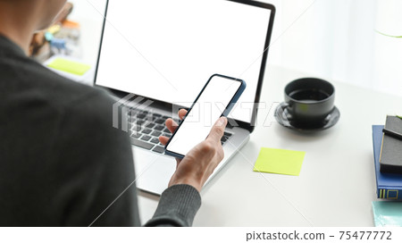 Over the shoulder view of young man freelancer using mobile phone and working with computer laptop at modern workplace. Over the shoulder view of young man freelancer using mobile phone and working with computer laptop at modern workplace. 75477772
