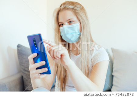 Young, blonde girl scrolling down in smartphone while wearing a blue medical face mask Young, blonde girl scrolling down in smartphone while wearing a blue medical face mask 75482078