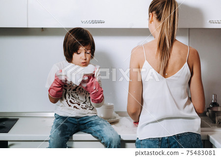 housewife mom in pink gloves washes dishes with her son by hand in the sink with detergent. A girl in white and a child with a cast cleans the house and washes dishes in homemade pink gloves.A child 75483081
