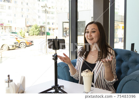 A young laughing brunette sits at a table in a cafe. She speaks by video communication on the phone, and on the table is a glass with a cocktail. She communicates via video link with a friend 75483396