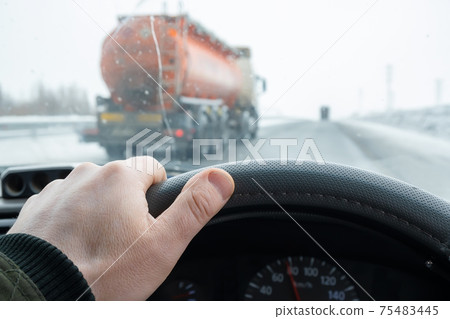 the driver's hand on the steering wheel of the car, which is overtaken on the secondary line by a tanker truck with a large tank of fuel 75483445
