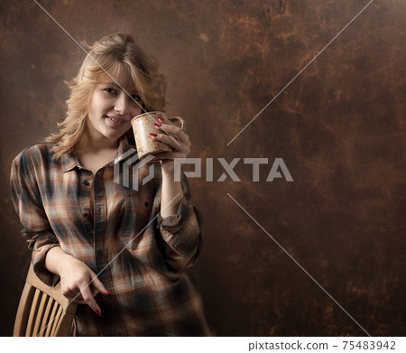 Pretty woman with long blonde hair in brown plaid shirt holds coffee or tea cup. Pretty woman with long blonde hair in brown plaid shirt holds coffee or tea cup. 75483942