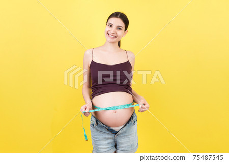 Cropped image of expecting mother in unzipped jeans measuring her pregnant belly with a tape measure at colorful background. Centimeter measurement 75487545