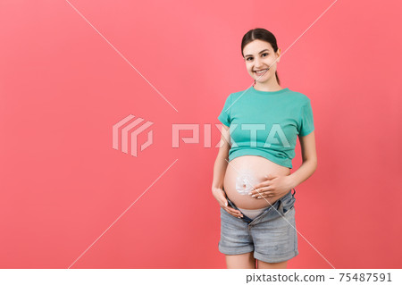 pregnant woman in unzipped jeans using moisture cream to prevent stretch marks on her belly at colorful background with copy space. Skincare concept pregnant woman in unzipped jeans using moisture cream to prevent stretch marks on her belly at colorful background with copy space. Skincare concept 75487591