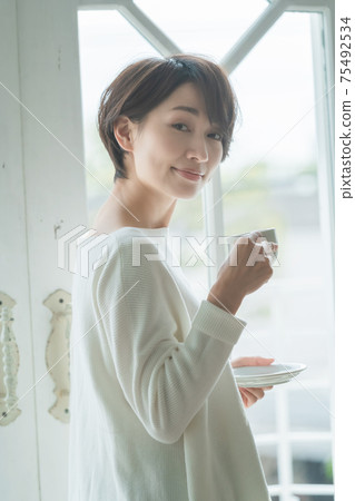 短頭髮的中量級家庭主婦站在窗戶旁邊,喝咖啡 短頭髮的中量級家庭主婦站在窗戶旁邊,喝咖啡 75492534