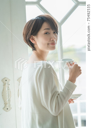 短頭髮的中量級家庭主婦站在窗戶旁邊，喝咖啡 75492535