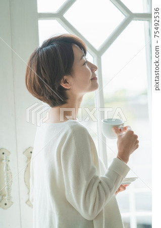 短頭髮的中量級家庭主婦站在窗戶旁邊,喝咖啡 短頭髮的中量級家庭主婦站在窗戶旁邊,喝咖啡 75492536