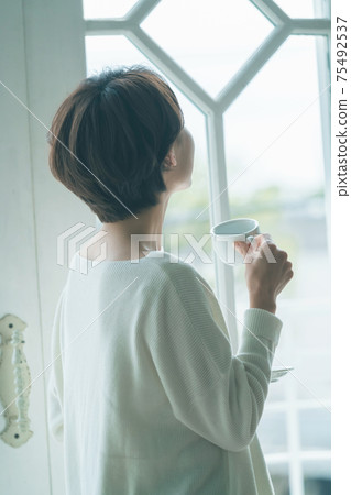短頭髮的中量級家庭主婦站在窗戶旁邊,喝咖啡 短頭髮的中量級家庭主婦站在窗戶旁邊,喝咖啡 75492537