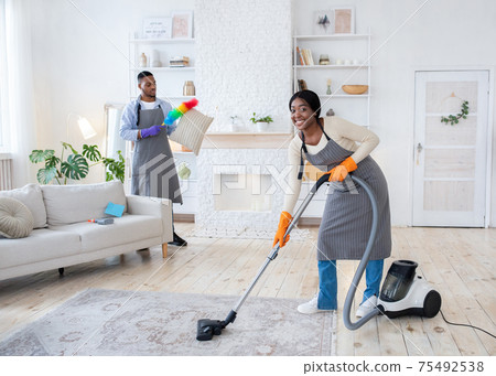 Happy black woman vacuuming floor while her boyfriend dusting cushion at their apartment, copy space Happy black woman vacuuming floor while her boyfriend dusting cushion at their apartment, copy space 75492538