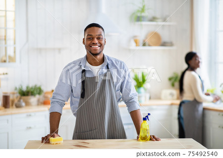 Portrait of black guy in apron and rubber gloves smiling at camera at kitchen, his wife washing dishes on background 75492540