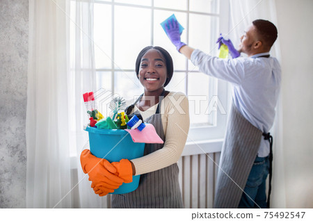 Cheerful black lady holding bucket of cleaning supplies while her husband wiping window at modern flat Cheerful black lady holding bucket of cleaning supplies while her husband wiping window at modern flat 75492547