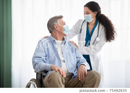 Young doctor and disabled senior man in wheelchair wearing face masks during covid-19 pandemic at retirement home Young doctor and disabled senior man in wheelchair wearing face masks during covid-19 pandemic at retirement home 75492635