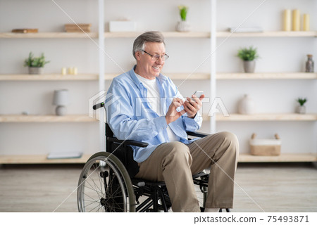 Positive senior impaired man in wheelchair checking messages or watching video on smartphone at home 75493871