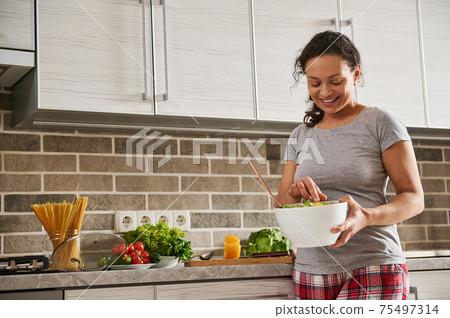 Smiling African American woman stirring vegetable salad with wooden spoon while smiling in kitchen. Houselife and healthy eating concept 75497314