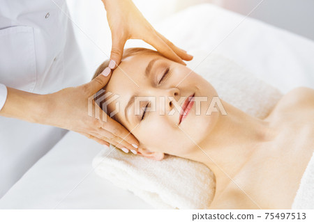 Beautiful caucasian woman enjoying facial massage with closed eyes in sunny spa salon. Relaxing treatment in medicine 75497513