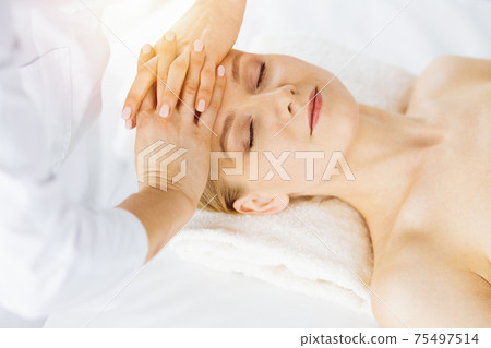 Beautiful caucasian woman enjoying facial massage with closed eyes in sunny spa salon. Relaxing treatment in medicine 75497514